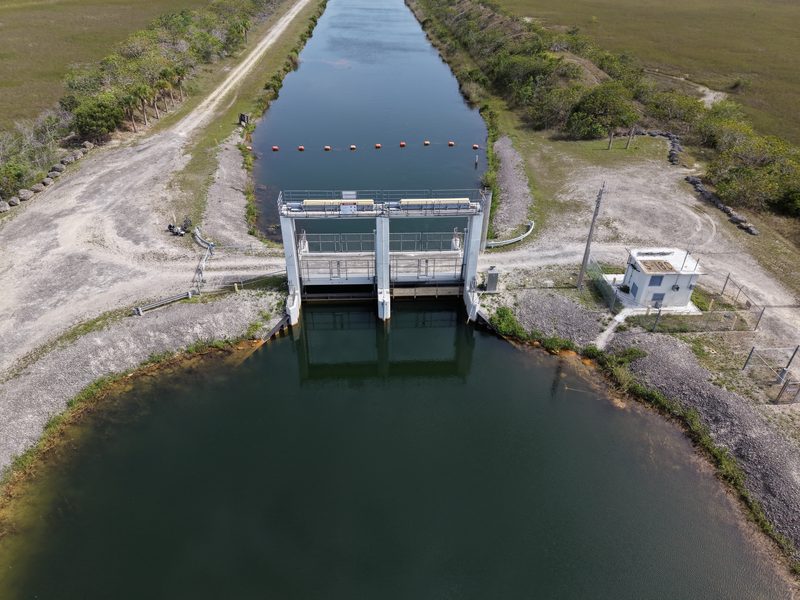 Wide aerial view of a water control structure on a canal between wetland tracts, with a small equipment building beside it.
